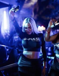 mujer rubia joven en una fiesta sostiene un vaper con un brazo levantado. Tiene una remera de g-pulse